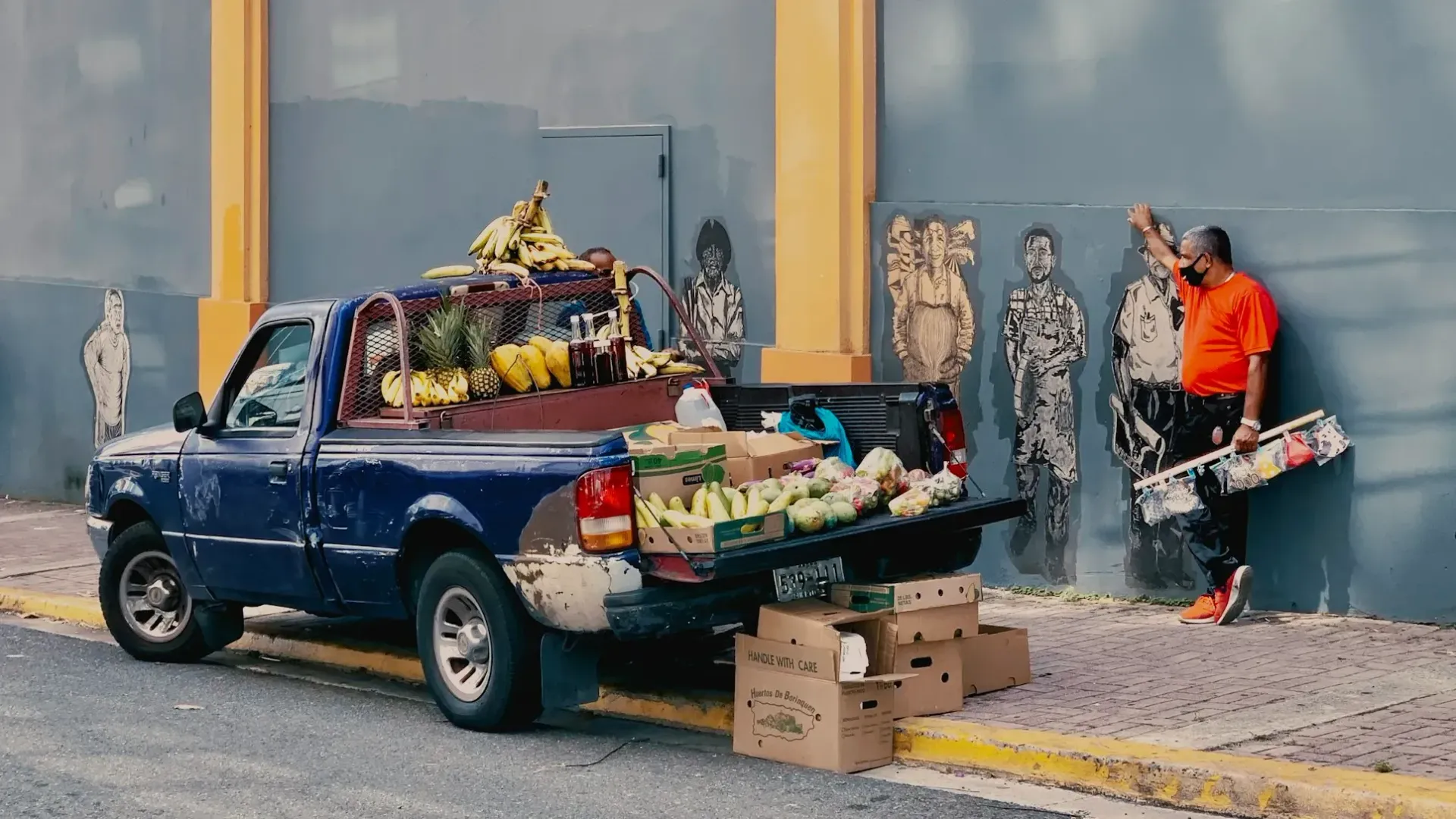 A man is standing next to a blue truck with fruit in the back.