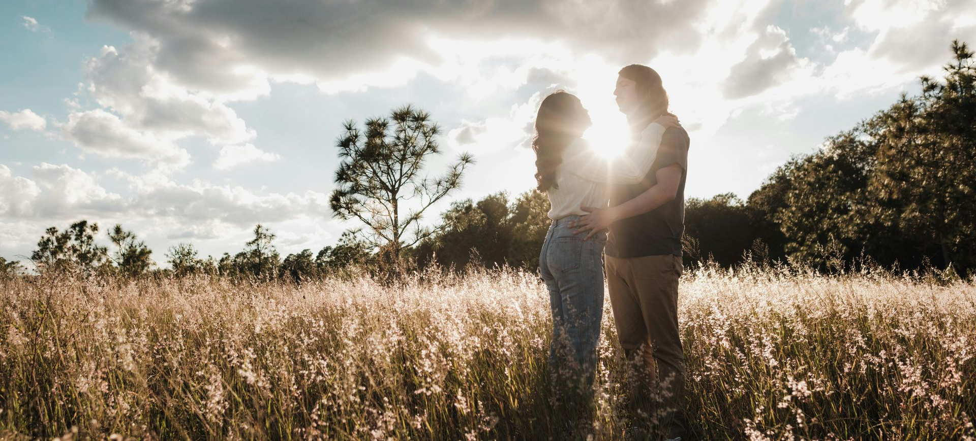 Couple embracing in a sunlit field, silhouetted. Sunlight glows between them, with tall grass in the foreground.