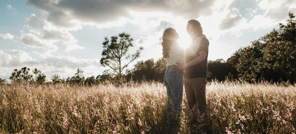 Couple embracing in a sunlit field, silhouetted. Sunlight glows between them, with tall grass in the foreground.
