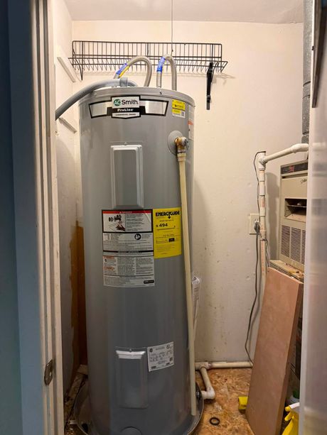 Gray water heater in a narrow utility closet with pipes and wires above.