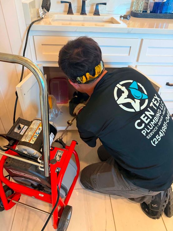Plumber kneels under a sink, working on a red drain-cleaning machine in a bathroom.