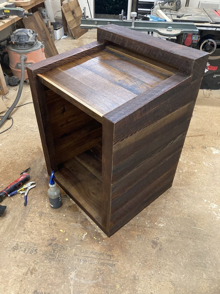 Dark wooden cabinet or stand under construction in a workshop, with tools on the floor nearby.