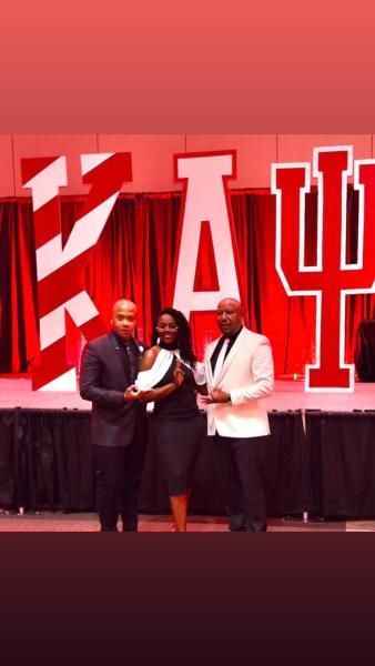 Three people on a red-lit stage holding an award in front of large red letters.