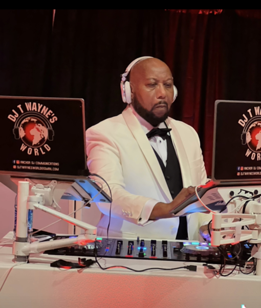 DJ in white tuxedo at a mixing console with laptops, performing under red stage lighting