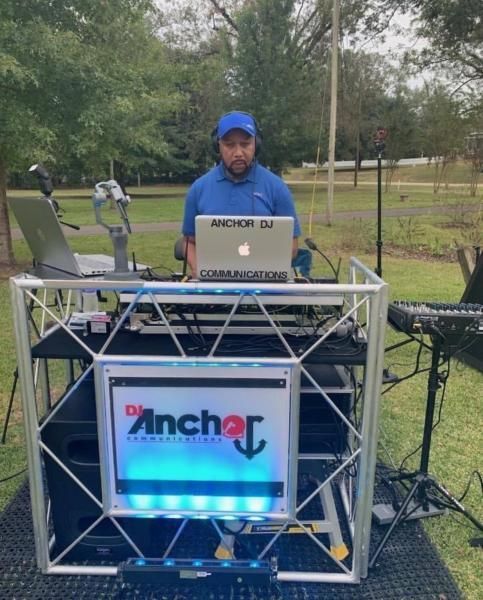 DJ at outdoor booth using laptop, wearing blue cap and shirt, with lit Anchor sign on front