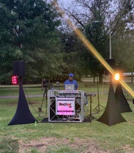 DJ at outdoor park setup with speakers and colorful lights behind a laptop booth