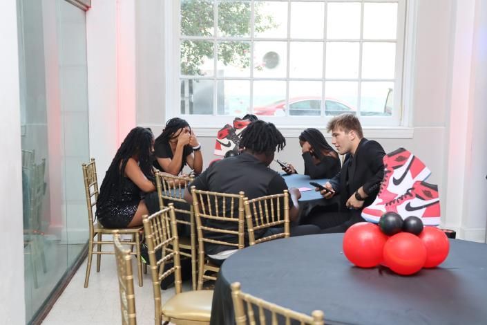 People seated at a café table with gold chairs near a window; red balloons sit on a nearby table.