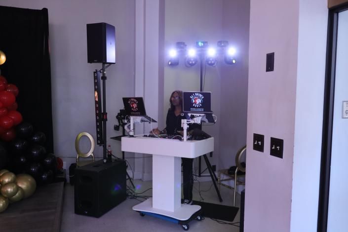 White DJ booth with laptop and lights in a small indoor event space, with speakers and balloons nearby