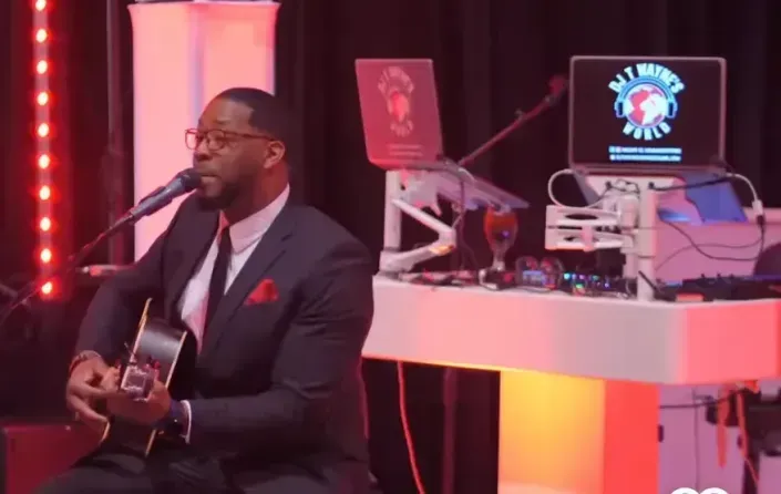 Man performing acoustic guitar on stage beside DJ equipment and glowing red lights