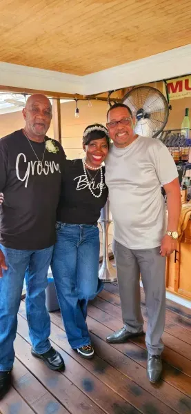 Three people smiling indoors, standing together on a wooden floor.