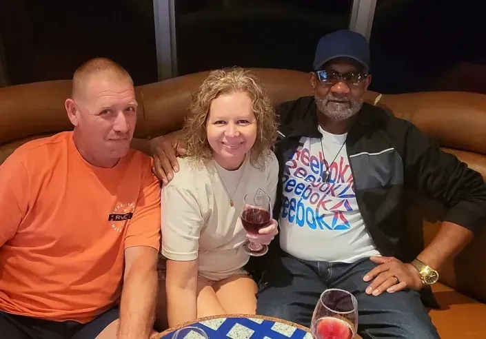 Three people smiling on a couch, holding drinks in a cozy lounge setting.