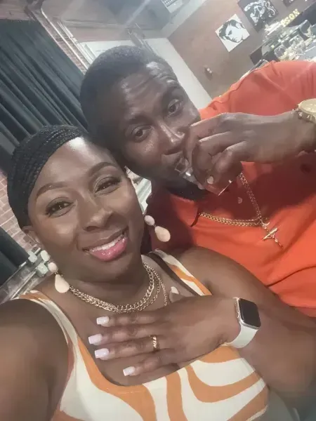 Two smiling people posing indoors, one in an orange shirt, the other in a striped top and smartwatch.