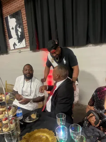 People seated at a table at a social gathering, with a man leaning over to speak to them.