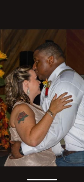 Two people embrace and kiss at a formal event, one in a white shirt and the other in a sleeveless dress.