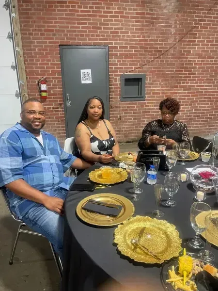 Three people seated at a dining table with plates, glasses, and food in a brick-walled room.