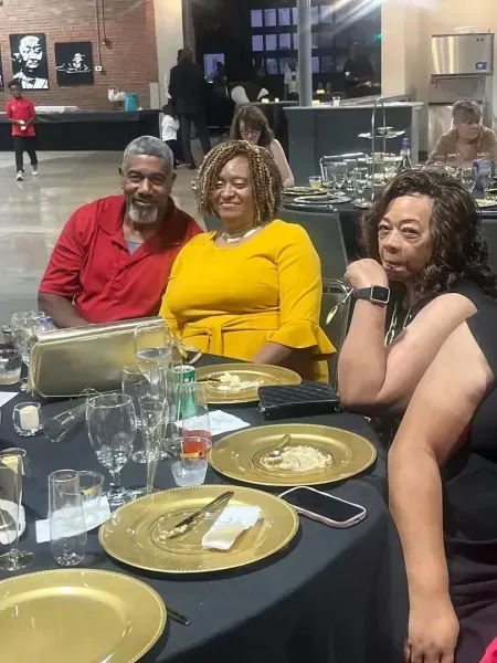 Three people seated at a restaurant table with plates and glasses, smiling for a photo.