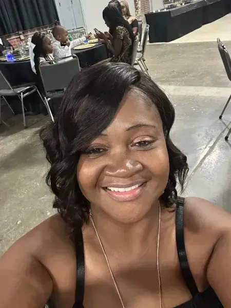 Smiling person taking a selfie at an indoor event, with tables and people in the background.