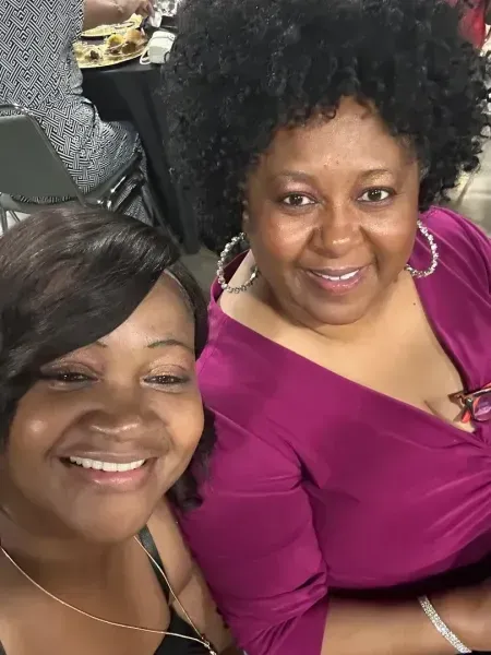 Two smiling women seated closely together, one in a magenta top, taking a selfie indoors.