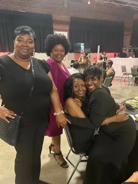 Four women pose and hug at an indoor event, with pink and black outfits and banquet tables in the background.