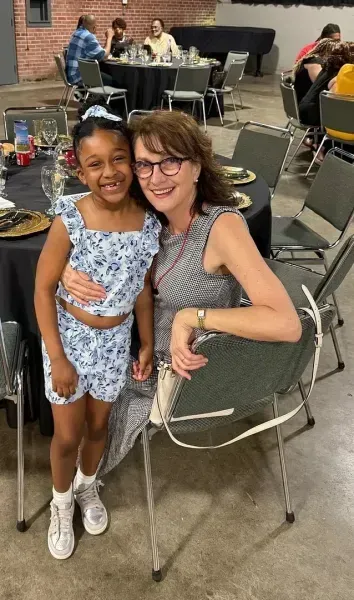 Two smiling people pose together indoors, seated at a table in a community hall.