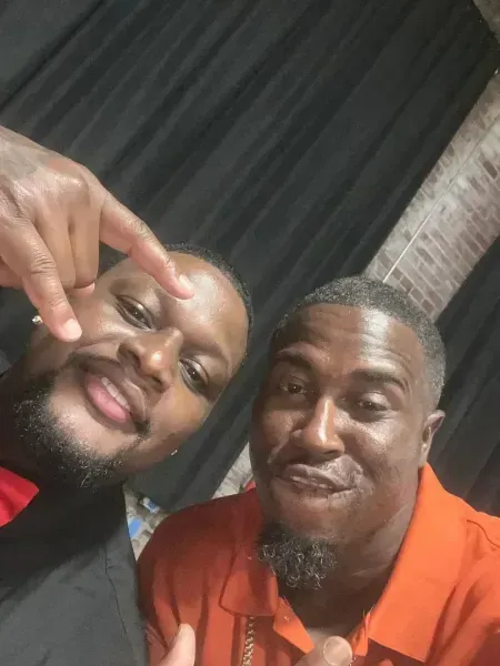 Two men smiling close together in a casual selfie, one making a hand gesture, indoors.