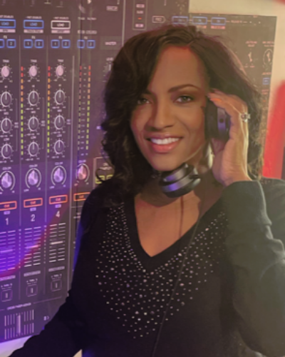 Smiling person wearing headphones in a dim studio, hand to ear, with audio mixer screens in the background