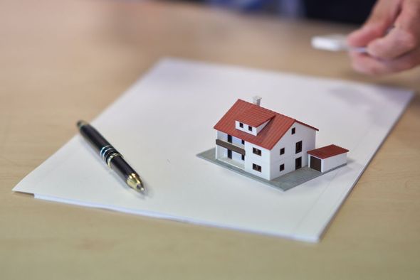 Modellino di una casa in miniatura su carta, disegnato con una penna nera su una scrivania; la mano è visibile sullo sfondo.