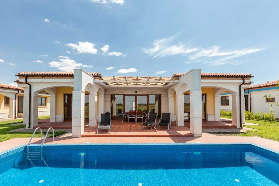 Patio di una villa moderna con lettini prendisole e una piscina blu sotto un cielo luminoso.