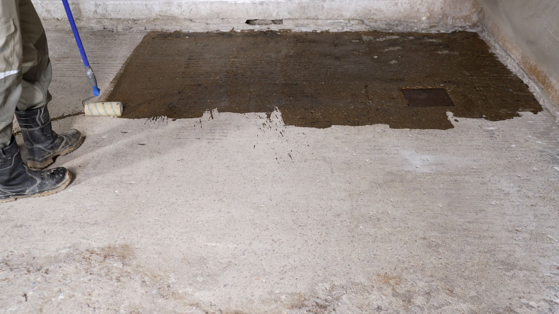 Partially finished concrete floor with a dark patch being spread near a wall, and a worker’s boots at left Partially finished concrete floor with a dark patch being spread near a wall, and a worker’s boots at left