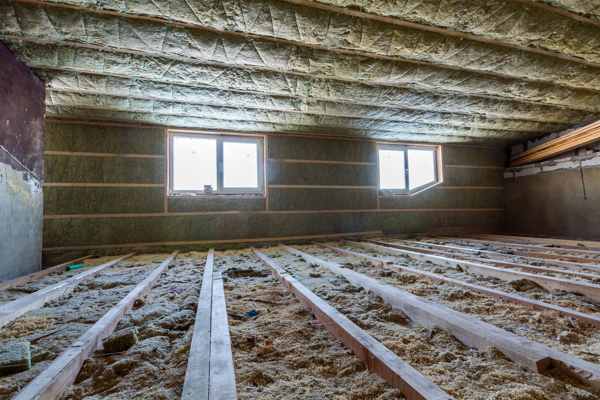Insulated attic with exposed floor joists and two small windows letting in daylight