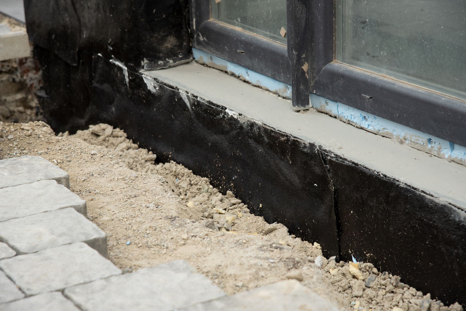 Black foundation wall beside a window, with gravel and stone pavers along the edge.
