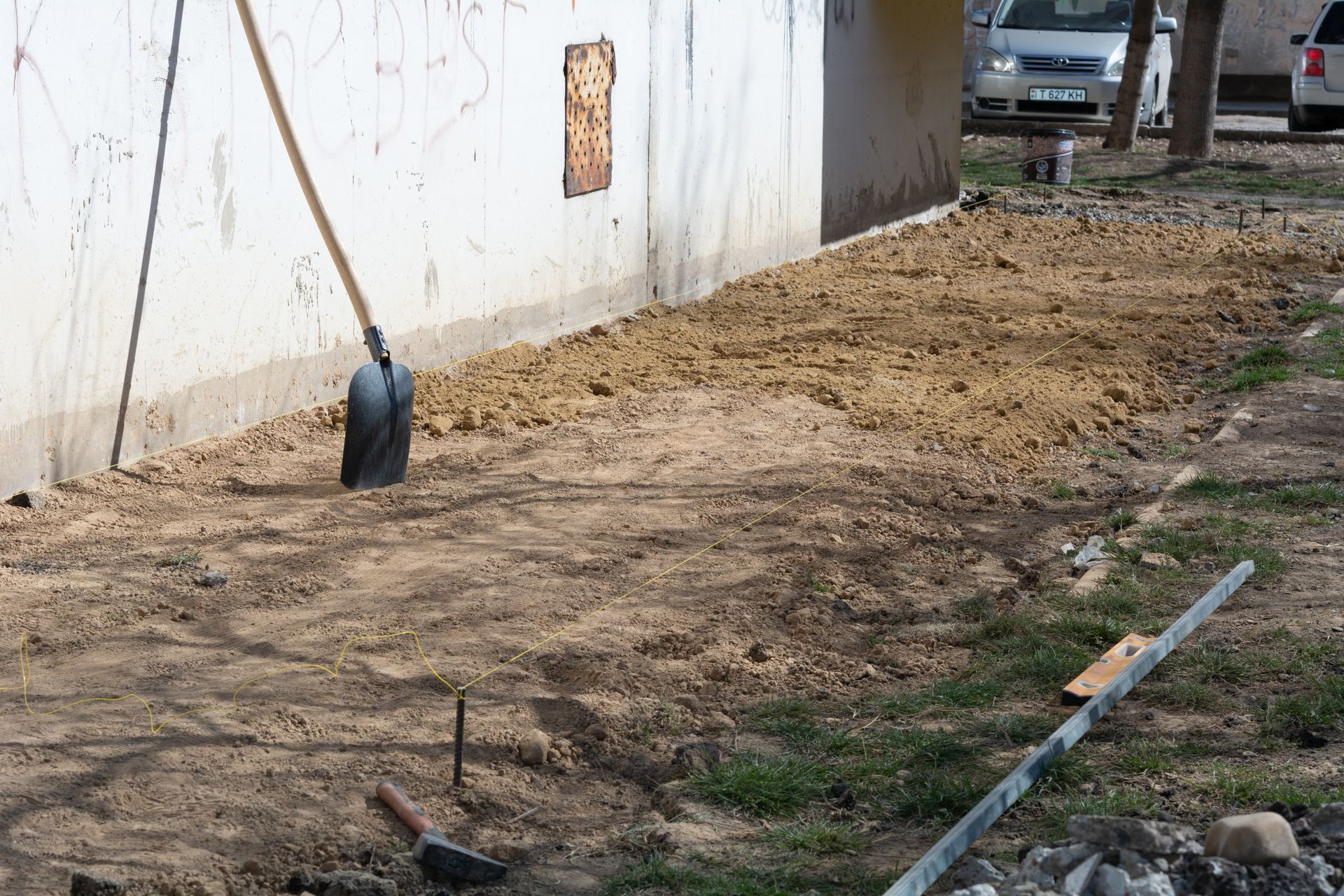 Construction tools beside a white wall on a dirt patch near a parked vehicle