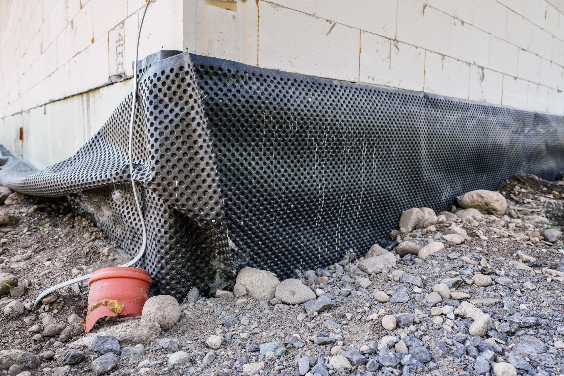 Black mesh erosion barrier over gravel beside a concrete wall, with a red pipe end visible.