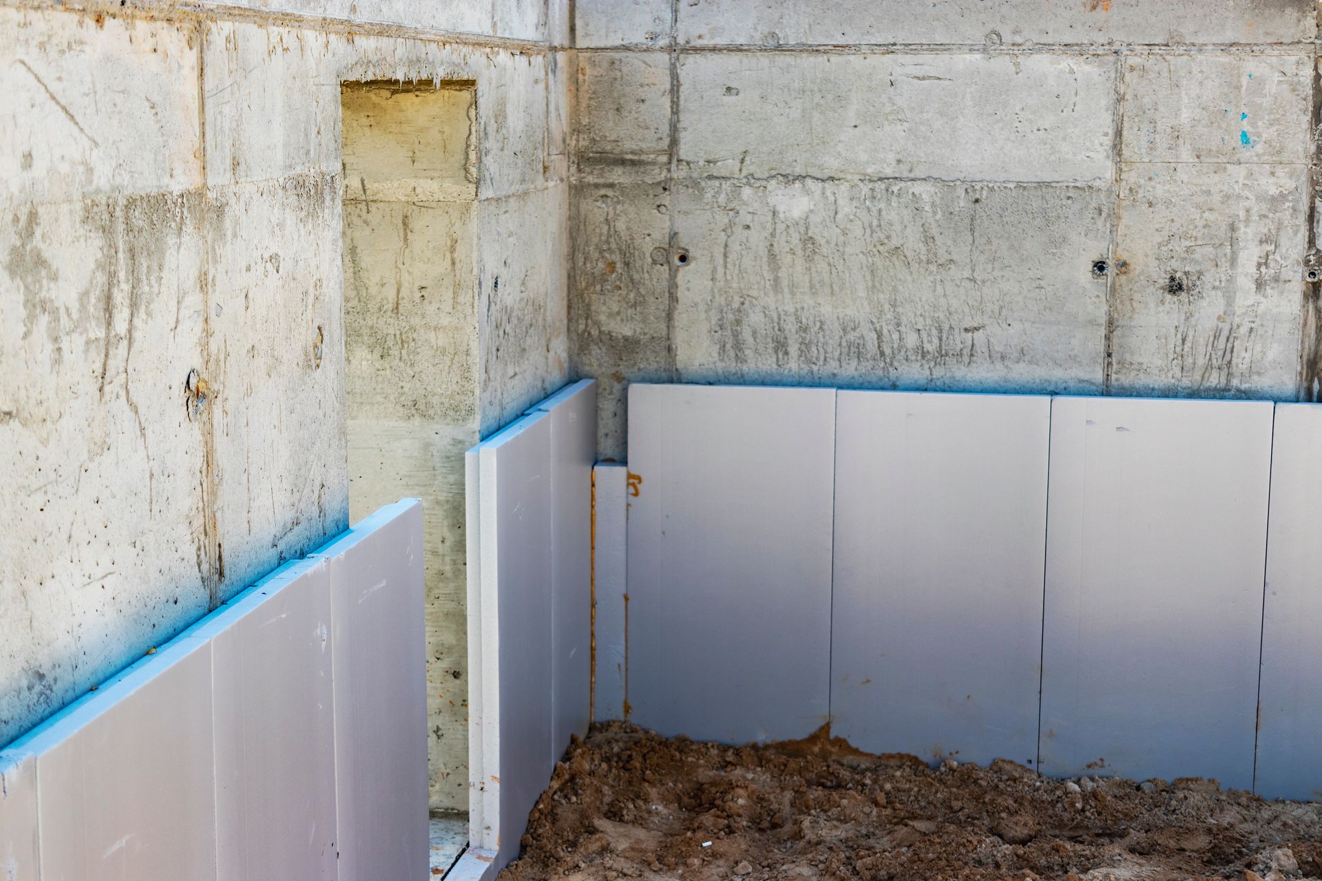 Concrete basement corner with blue-topped wall insulation panels and dirt floor