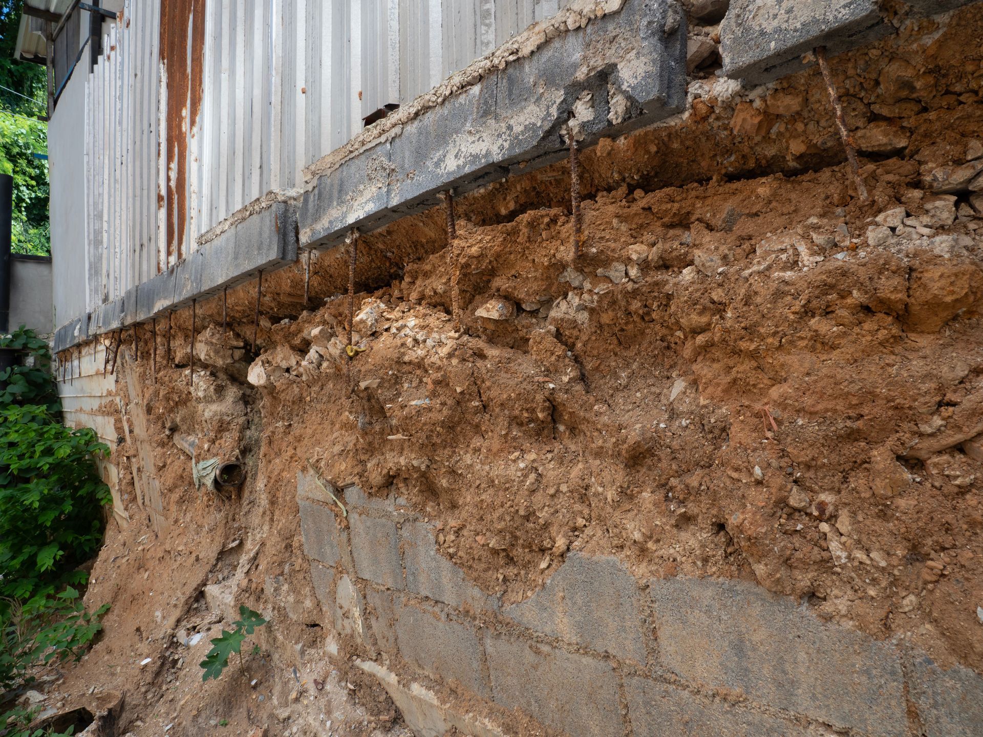 Eroded dirt slope beneath a corrugated metal wall and concrete blocks, with exposed soil and greenery nearby Eroded dirt slope beneath a corrugated metal wall and concrete blocks, with exposed soil and greenery nearby