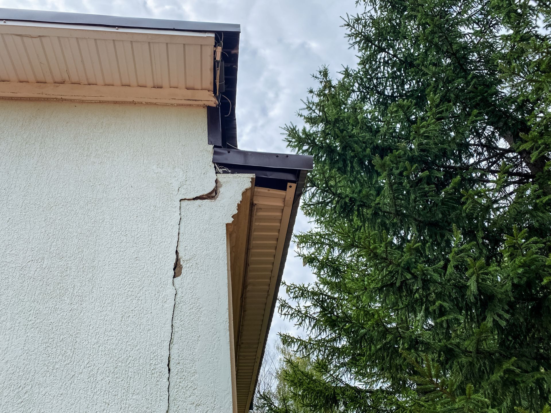 Cracked stucco exterior wall under a roof eave beside a tall evergreen tree Cracked stucco exterior wall under a roof eave beside a tall evergreen tree