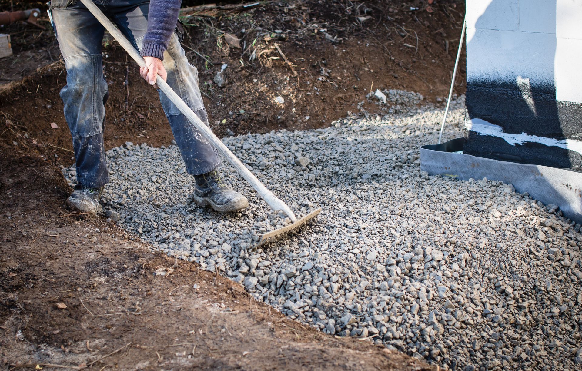 Person raking gravel on a construction site beside a dirt path and foundation wall