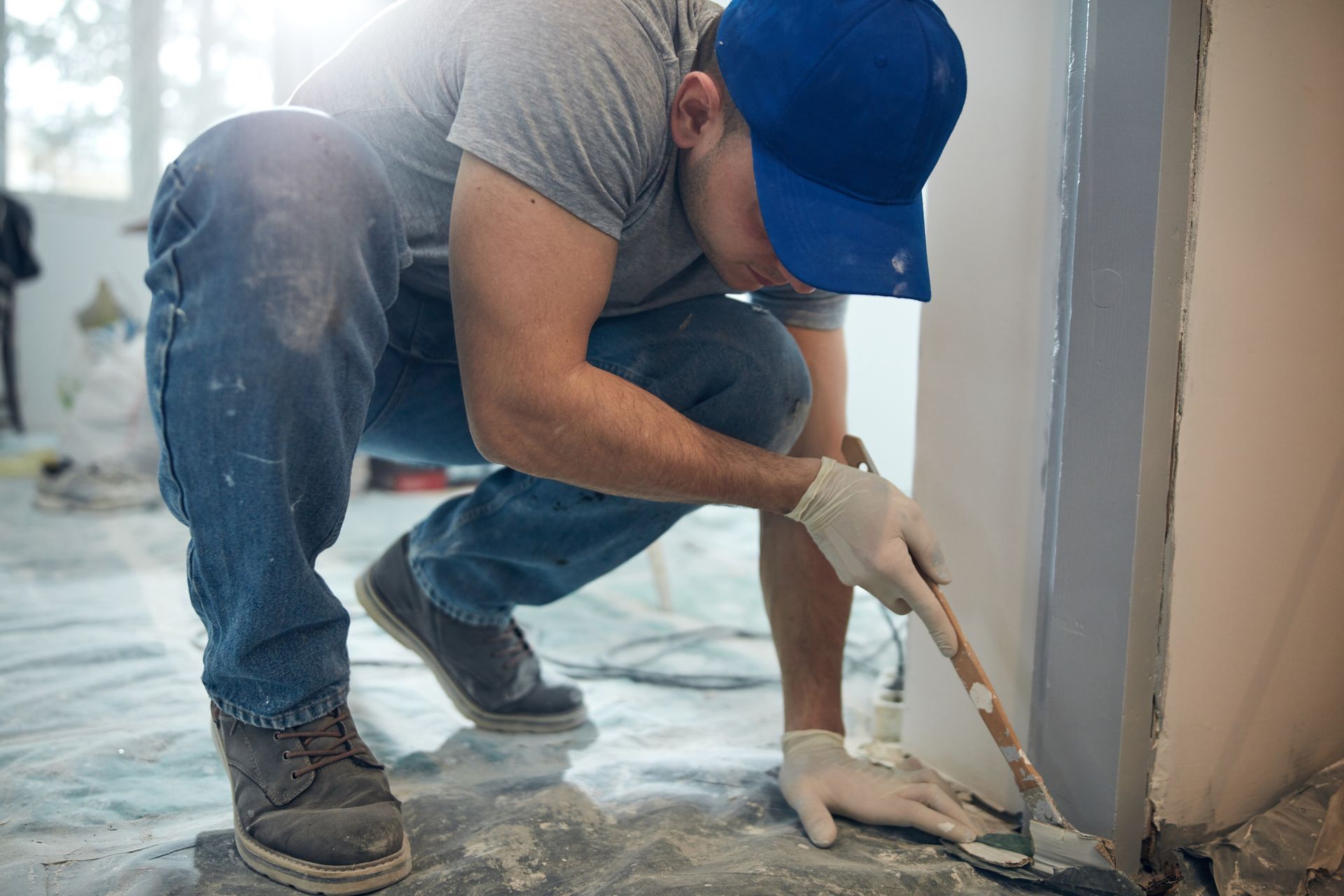 Worker in a blue cap scrapes plaster from a wall during home renovation.