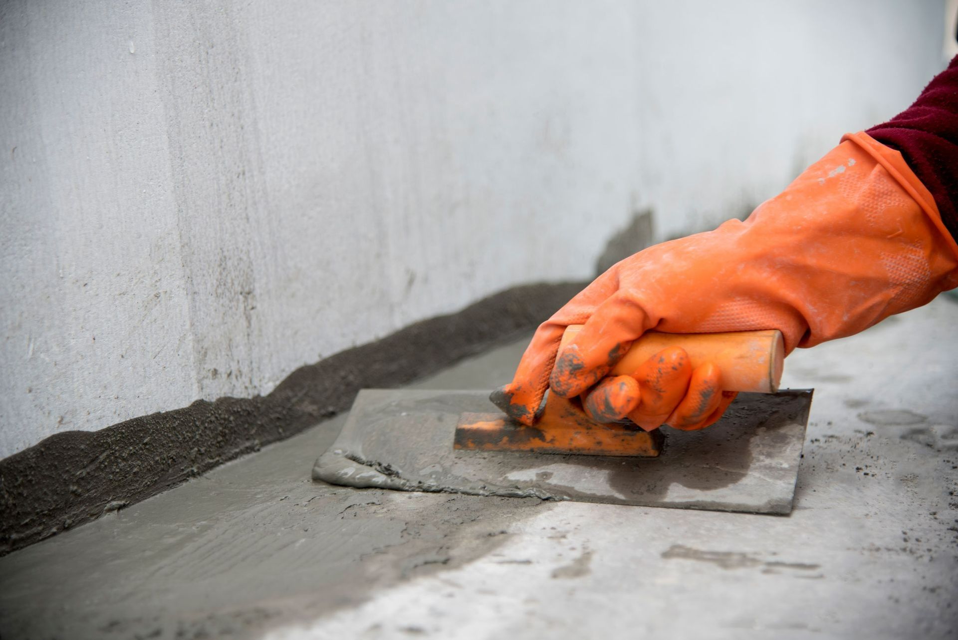 Orange-gloved hands trowel wet concrete along a wall indoors Orange-gloved hands trowel wet concrete along a wall indoors