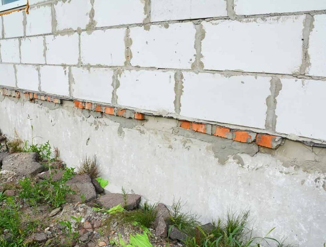 Partially built cinder block wall with exposed orange bricks and weeds at the base