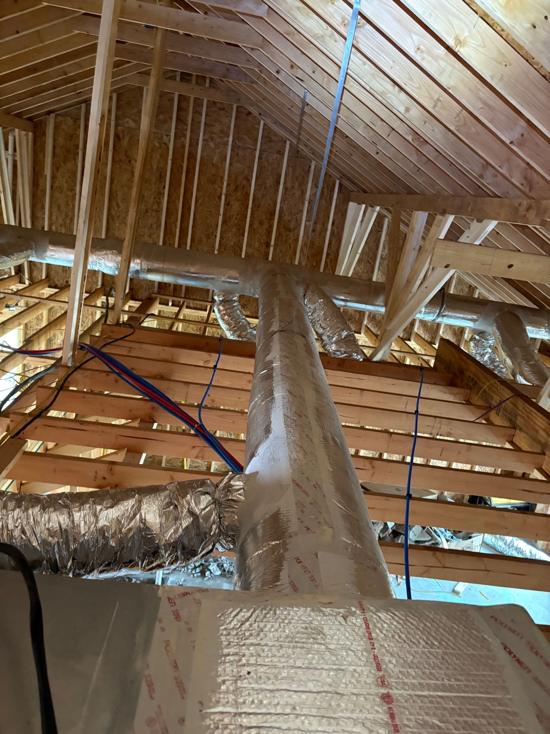 Inside an attic, exposed wooden rafters and beams with silver insulation and a blue cable hanging down.