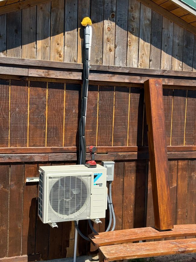 Outdoor air conditioner unit mounted on a rustic wooden wall beneath a black vent pipe