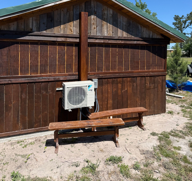Wooden shed with a wall-mounted air conditioner unit and a bench in front on a dirt ground.