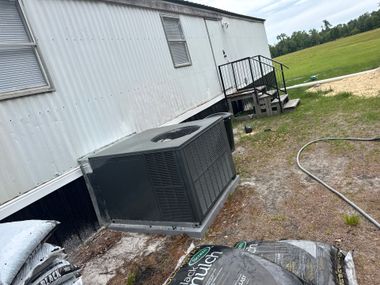 Outdoor HVAC unit beside a white mobile home, with a gravel yard and grassy field in the background.