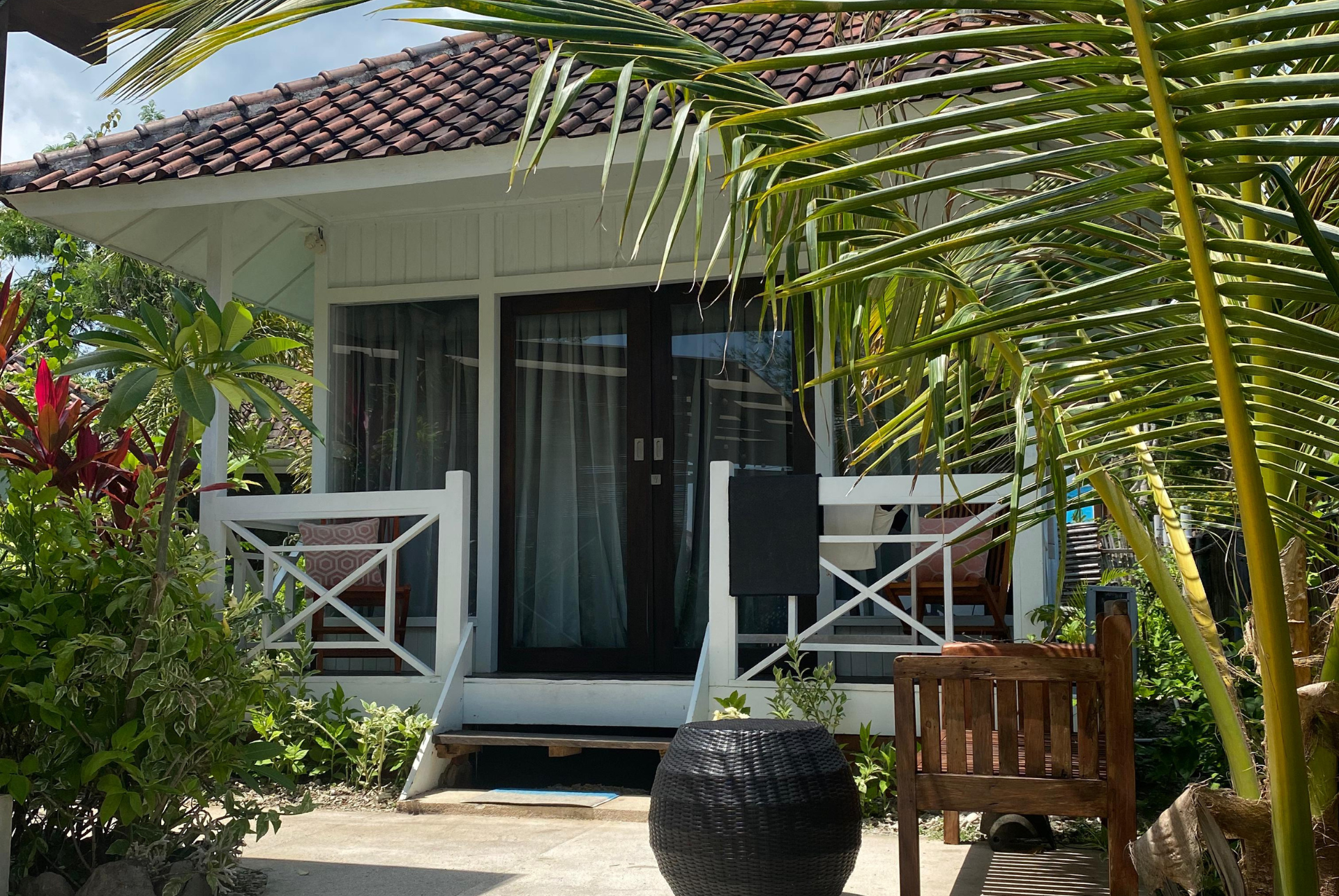 White bungalow with porch, brown tile roof, surrounded by tropical foliage.