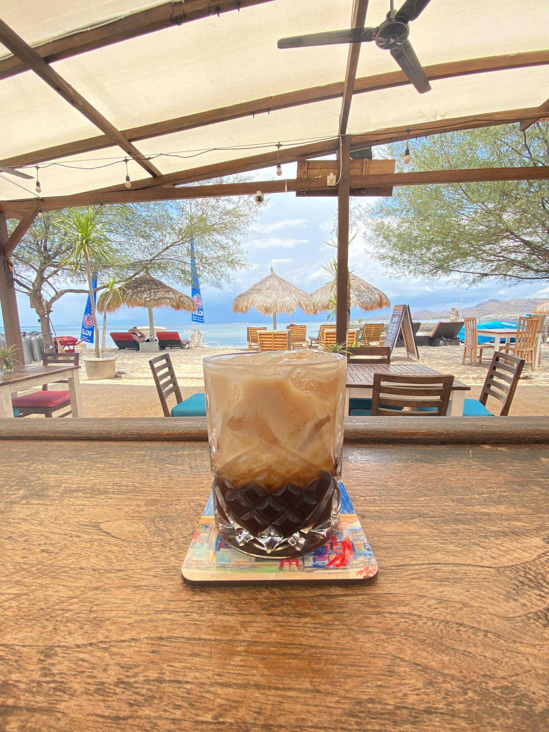 A glass of iced coffee is sitting on a wooden table in front of a beach.
