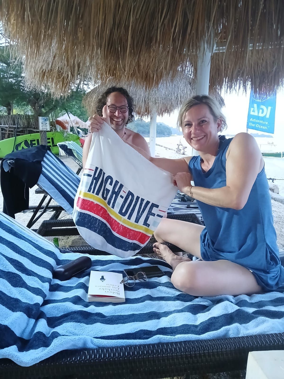 Two women are sitting on a beach holding a towel that says high dive