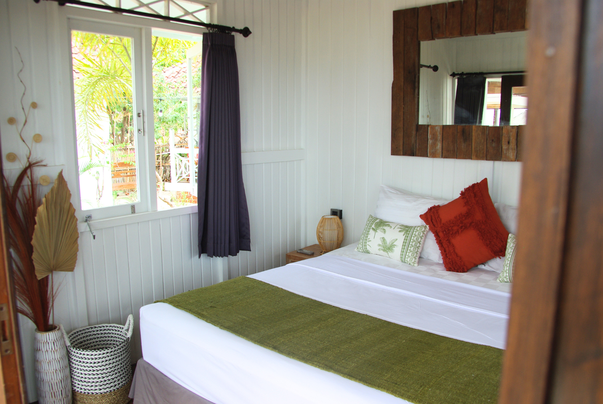 Bedroom with white walls, bed with green runner, orange pillow, wood-framed mirror, and window with purple curtain.