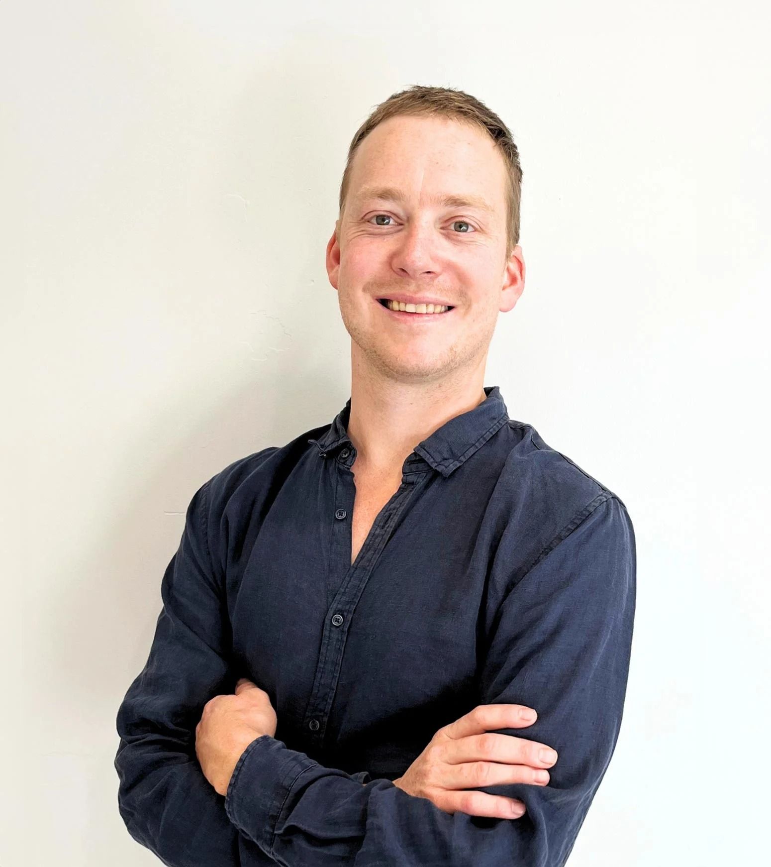 Smiling man in a dark blue shirt standing with arms crossed against a white wall