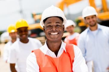 A smiling construction worker wearing a white hard hat and orange safety vest stands in front of a blurred work crew.